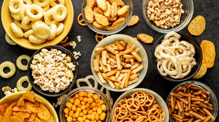 Assorted chips, popcorn, onion rings, croutons, cheese balls, biscuits in bowls. unhealthy food for figure, heart, skin, teeth. An assortment of fast carbohydrates. Junk food on a stone background