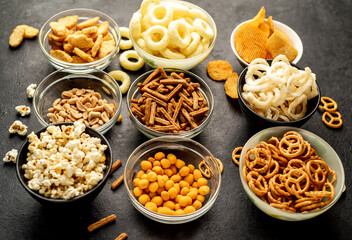 Assorted chips, popcorn, onion rings, croutons, cheese balls, biscuits in bowls. unhealthy food for figure, heart, skin, teeth. An assortment of fast carbohydrates. Junk food on a stone background