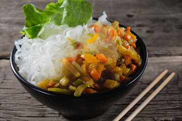 A plate of rice noodles stands on an old wooden table top. The side dish consists of fried onions, carrots, paprika.