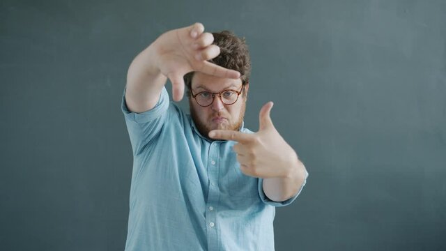 Slow motion portrait of creative guy making focus hand gesture and smiling looking at camera standing on gray background searching beautiful view.