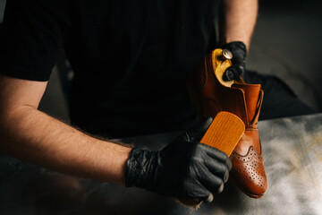Close-up view of shoemaker in black gloves polishing light brown leather shoes with brush during restoration working. Concept of cobbler artisan repairing and restoration work in shoe repair shop.