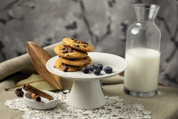 Horizontal capture of chocolate chip cookies lined upwards with bottle of milk, blueberries, cinnamon bark in wooden plate and lace ground in frame