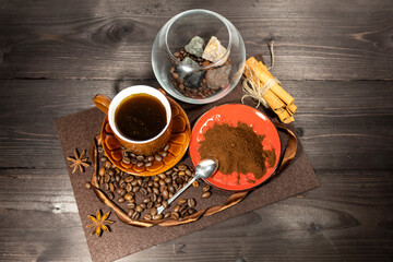 There is a cup of coffee on the table, ground coffee on a red saucer. Scattered nearby are coffee beans, cinnamon sticks and anise stars. In the background, coffee beans and gems lie in a glass vase.