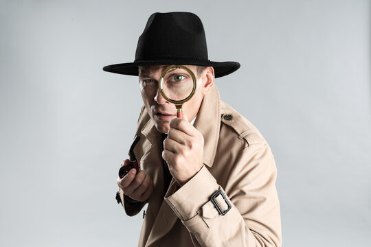 Male detective with smoking pipe looking through magnifying glass on grey background