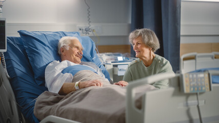 Hospital Ward: Elderly Man Resting in Bed, His Caring Beautiful Wife Supports Him Sitting Beside, Happy Couple Together, They Talk, Remember Good Times. Old Man Recovering successfuly After Sickness - obrazy, fototapety, plakaty