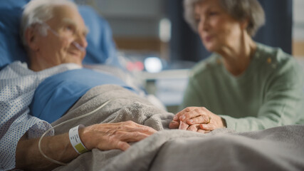 Hospital Ward: Focus on Hands of Elderly Man Resting in Bed, His Caring Wife Supports Him By...