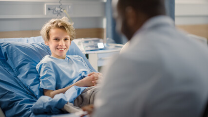 Hospital Ward: Handsome Young Boy Resting in Bed, Carrying Doctor Visits to Support Him, Doctor Talks, Gives Advice. Happy Smiling Patient Recovering after Sickness or Successful Surgery