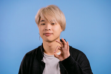 Asian man with dyed hair showing okay signs over blue background. A positive young korean guy smiles to camera. Winner. Success. Body language.