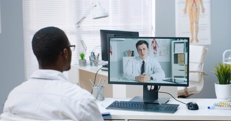 Close up of African American male physician speaking on online video conference on computer with Caucasian young coworker sitting in hospital cabinet, doctor consulting nurse on treatment on web call