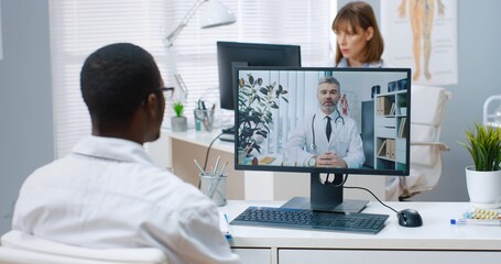 Close up. Over shoulder view of African American man doctor sitting in cabinet in clinic speaking with Caucasian young coworker consulting online on computer, medical web conference healthcare concept