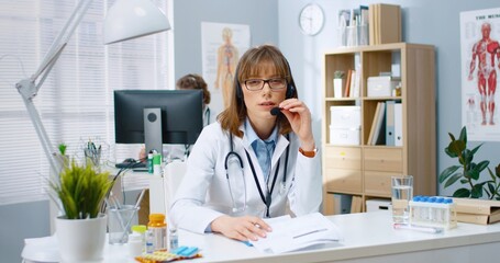 Portrait of beautiful young Caucasian female physician sitting in hospital at desk speaking and...