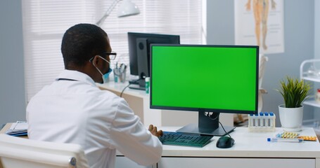 Back view of African American male physician sitting in hospital office and looking at green screen...
