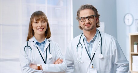 Portrait of Caucasian joyful team of doctors male and female colleagues in white coats standing in cabinet in clinic and smiling to camera. Professional physicians looking at camera, hospital concept