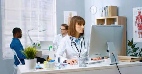 Caucasian young female physician working in medical center sitting at desk typing on computer at workplace while male colleague speaking with African American patient on background, medic concept