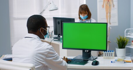 Back view of African American male physician sitting in hospital office and looking at green screen while working typing on computer. Medical concept, doctor browsing on monitor with chroma key
