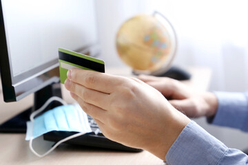 Fototapeta premium Man holding credit bank card types on PC keyboard. Concept of online shopping and payment, buying a travel tour during coronavirus pandemic