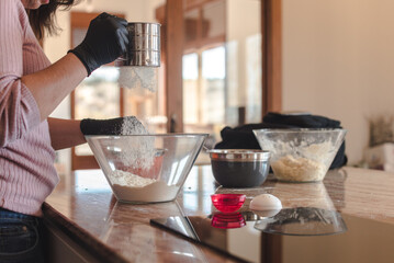 Mujer joven con guantes en su cocina mientras tamiza la harina en un recipiente de cristal. Concepto de cocinar pizza casera. Hacer pan en casa. - obrazy, fototapety, plakaty