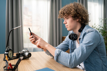 Portrait of a Caucasian Teenage Boy with Curly Hair in Cozy Living Room Using Smartphone at Home. He is Browsing the Internet and Checking Videos on Social Networks and Typing message.