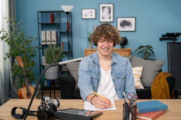 Smiling American millennial male student in headphones sit at desk think about webinar making...