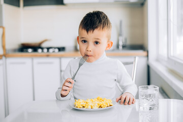 A child in the afternoon on a white light kitchen in a white sweater eats an omelet
