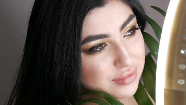 Very beautiful young woman model looks through green palm leaves posing in photo studio. Portrait of a girl with a bright evening smoky eye make-up, dark hair and brown eyes looks