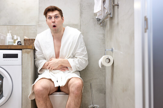 Man in bathrobe suffering from stomach ache on toilet bowl in bathroom