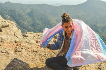 Cute Transgender Man Smiling Showing LGTB blue and pink Flag as Cape Sitting on the top of a Mountain