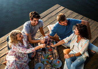 Friends having fun on picnic near a lake, sitting on pier eating and drinking wine.	