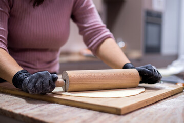 Una mujer amasa una masa de pizza con un rodillo sobre una tabla de madera. La cocinera lleva guantes negros. 