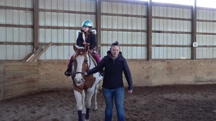 Female Instructor Teaching Young Girl How To Ride A Horse In An Indoor Riding Arena. wide shot