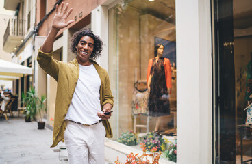 Smiling hipster guy in casual wear walking with smartphone enjoying free time and traveling on urban settings, positive curly handsome man waving using mobile phone for sending mails via 4G connection