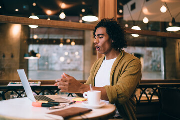 Handsome emotional 20s hipster guy watching videos on laptop computer sitting in cafe interior,expressive young millennial man surprised with news from social networks pointing on monitor