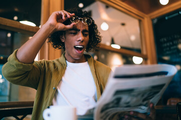 Emotional curly hipster guy amazed with latest news from printed media taking off eyewear while reading, expressive male 20s shocked with financial economical information about business on leisure
