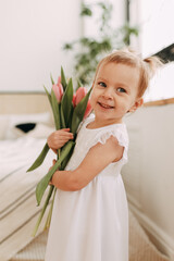 Cute cheerful smiling happy little girl child in a white dress holding a bouquet of pink tulip flowers laughs rejoices and looks