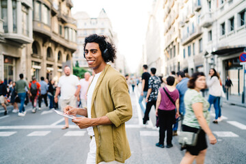 Half length portrait of funny male blogger in electronic earphones crossing city road and smiling at camera during vacations journey, happy hipster guy with digital tablet listening online audio guide