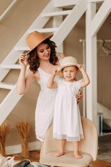 Happy beautiful mother and daughter in white dresses and straw hats try on fashionable outfits have fun laugh and pose together for the camera in the room of the house prepare for the summer vacation