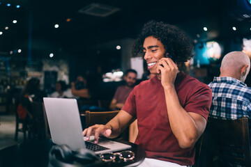 Happy male watching funny video content and smiling while discussing social media during smartphone conversation in coffee shop, cheerful freelancer browsing network website on laptop and calling