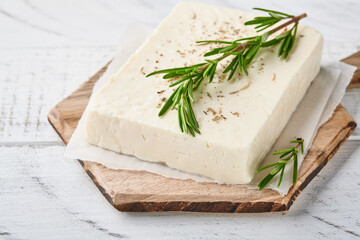 Cheese feta with rosemary, herbs and olive oil on wooden cutting board on white old wooden background. Traditional Greek homemade cheese. Selective focus.
