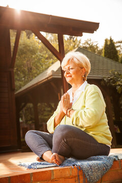 Active senior woman meditate in her garden.She sitting in lotus position in front of her home.