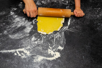 one female hand together with child’s hands roll a rolling pin over pastry or dough to flatten or shape it on a table covered with flour. Cooking together with a child at home. Top view.