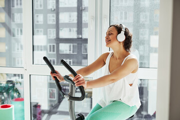 Young attractive woman listen to the music and working out on spin bike against large windows at...