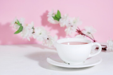 Fototapeta na wymiar White mug with pink tea, on a saucer, on a concrete table, on a pink background with flowers. Hard shadows. The drinks. Copy space