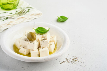 Feta cheese cubes with rosemary, olives and olive oil sauce in white bowl on light gray background. Traditional Greek homemade cheese. Selective focus.
