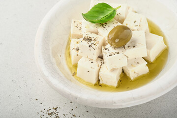 Feta cheese cubes with rosemary, olives and olive oil sauce in white bowl on light gray background. Traditional Greek homemade cheese. Selective focus.