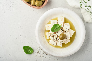 Feta cheese cubes with rosemary, olives and olive oil sauce in white bowl on light gray background. Traditional Greek homemade cheese. Selective focus.