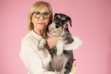 Senior woman holding her schnauzer puppy over pink background.