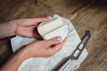 Cut Camembert cheese, French soft cheese with white mold in the hands of a woman on the background of a craft textile tablecloth, close-up. Knife for slicing cheese