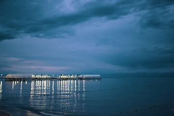 Obraz premium Twilight landscape of pier stretching out into sea. Illumination and umbrellas on ocean bridge. Glare from lamps on water. Nature landscape. Tourism background. Travel and vacation. Cloudy sky.