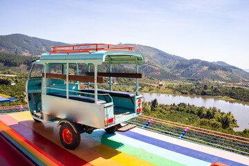 a vintage old decorated blue car with an open trunk and red wheels above a rainbow colored ground on a background of high green mountains and farms with a lake