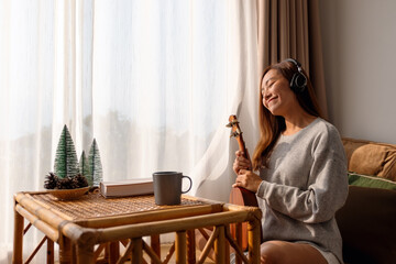 A beautiful young asian woman holding an ukulele and enjoy listening to music with headphone at home